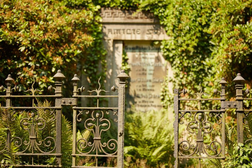 alter, gusseisener Zaun auf dem Friedhof, dahinter alte Grabstätte und Bäume