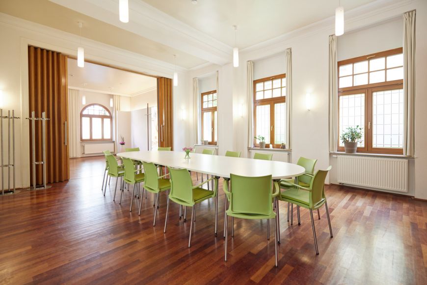 Gemeindesaal im Gemeindehaus: heller Raum mit dunklem Parkett-Boden, in der Mitte große Tafel mit vielen Stühlen, große Fenster