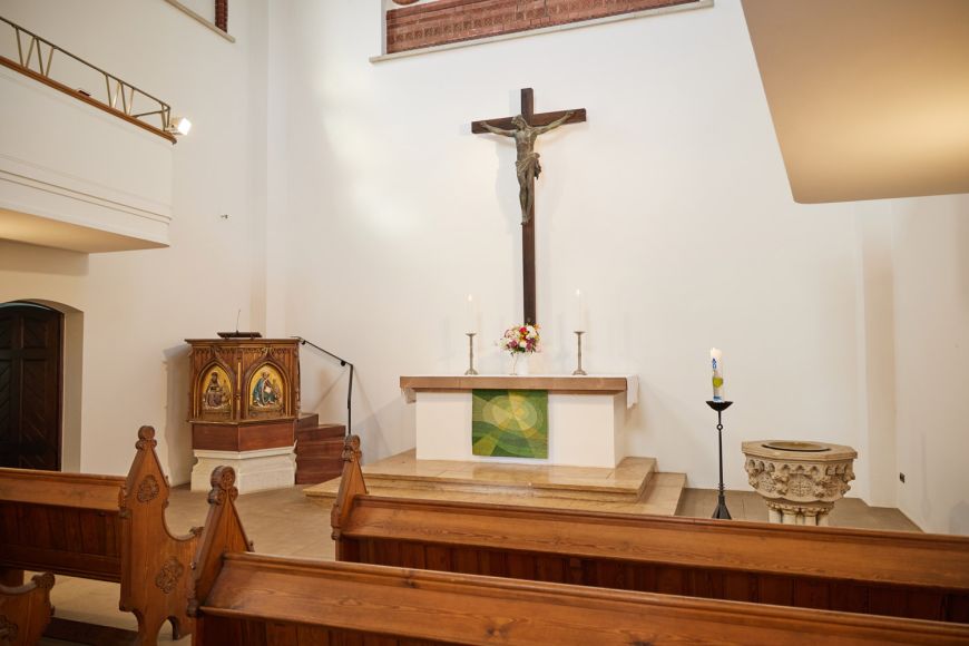 Innenraum der Kirche mit Holzbänken, steinernem Altar, Kanzel, Taufbecken und Kruzifix