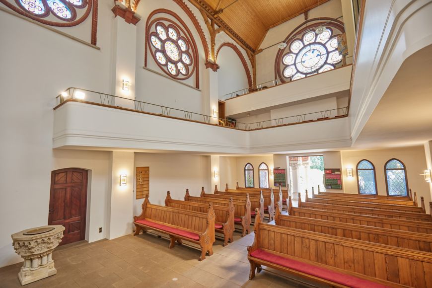 Innenansicht Kirche, Blick nach hinten Richtung Eingang. Hölzernes Gestühl, mittig Gang zum Eingang. U-förmige Empore über dem Gestühl, darüber Orgelempore. Auf deren Höhe drei große, bunte Fenster in Form von Blumen.