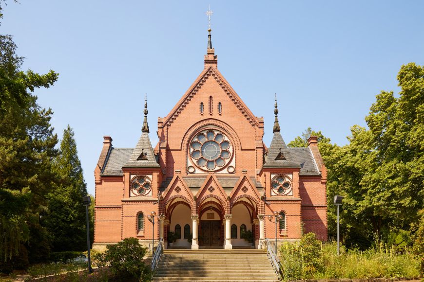 Außenansicht Kirche: kompakter Backsteinbau, Eingangsbereich mit Gewölbe und Ziertürmen. Zentrales Kirchenfenster in Blumenform über dem Eingang.