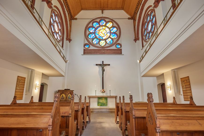 Innenansicht Kirchenschiff: hölzernes Gestühl, mittig Gang zum Alter. Darüber Kruzifix und großes, zentrales Kirchenfenster in Form einer Blume. Links und rechts Emporen, über ihnen nochmals bunte Fenster in Blumenform.