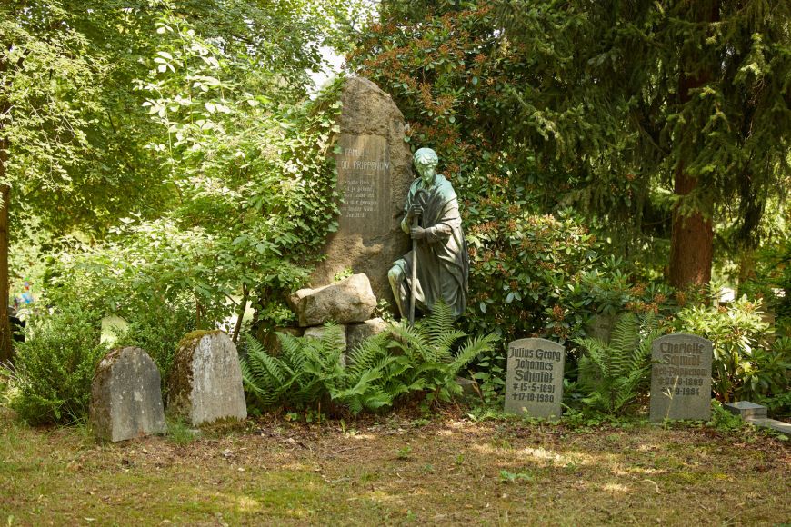 alte Grabsteine auf Friedhof, zentral Grabmal mit menschengroßer Statue, umgeben von Bäumen.